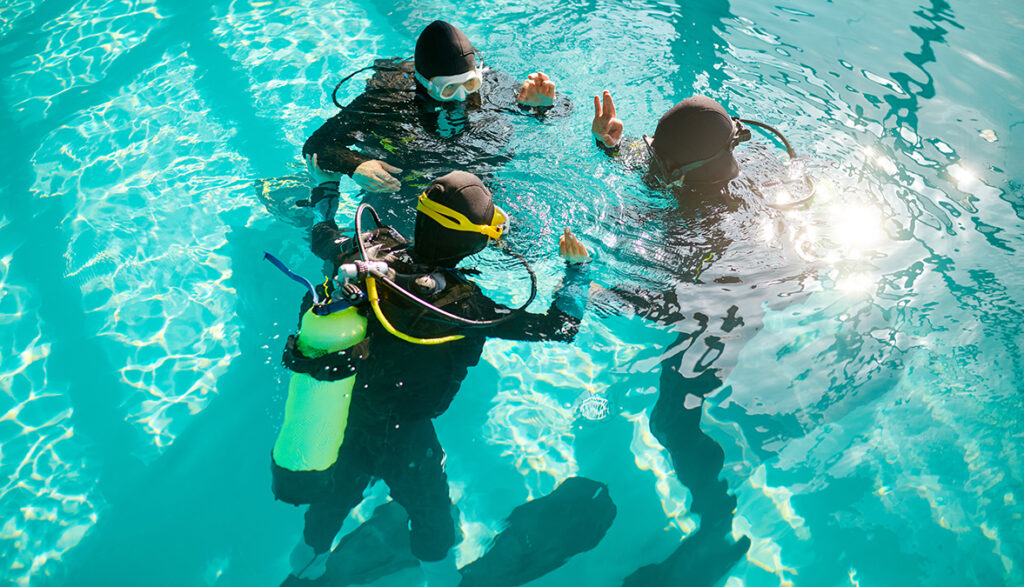 Divemaster and two divers in aqualungs, course in diving school. Teaching people to swim underwater with scuba gear, indoor swimming pool interior on background, group training