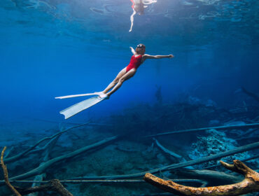 Freediver woman swims underwater with fins over sunken logs in the freshwater.