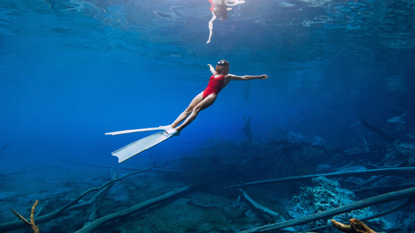 Freediver woman swims underwater with fins over sunken logs in the freshwater.