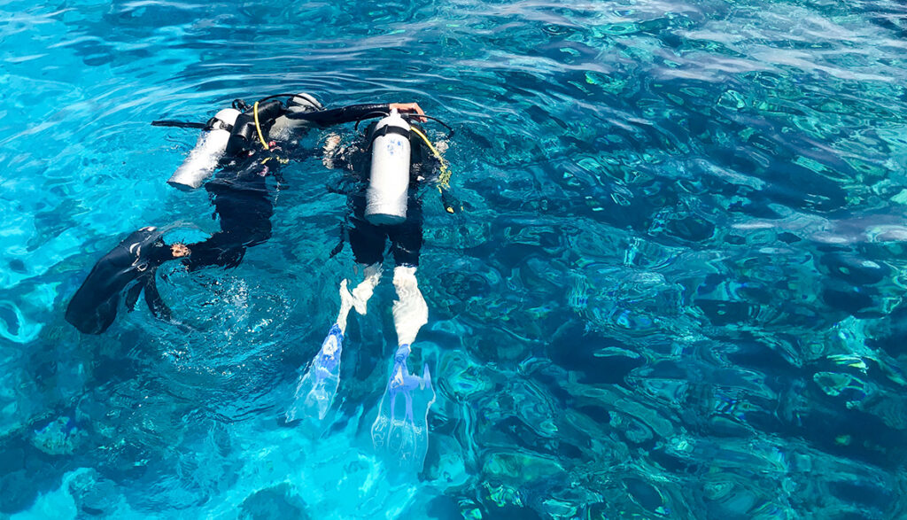 Two divers in black scuba diving suits, a man and a woman with oxygen bottles sink under the transparent blue water in the sea, the ocean in a tropical paradise warm resort