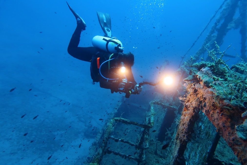 Sunken wreck and scuba diver. Travel photo, diver and shipwreck. Deep blue ocean with underwater photographer taking video of rusty metal shipwreck. Scuba diving trip, wreck exploration.
