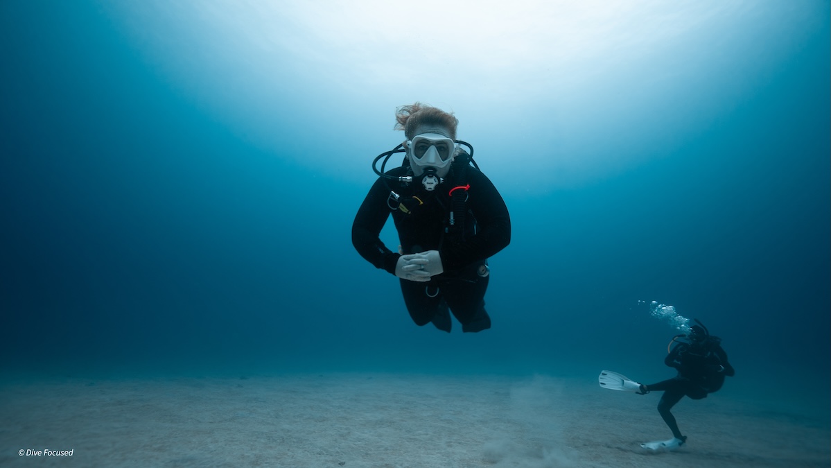 An underwater photograph of a scuba diver wearing a black wetsuit and a white mask, facing the camera with hands clasped together. The diver is hovering above a light sandy bottom in clear blue water. A second diver is swimming in the background to the right.