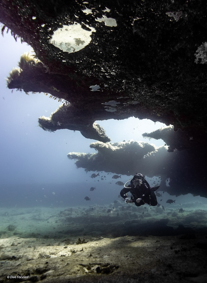 An underwater photograph of a scuba diver swimming horizontally beneath a large, dark, textured rocky overhang. Sunlight filters down past the edge of the reef, illuminating the diver and a school of small fish scattered in the water above a sandy seabed.