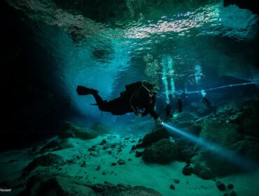 An underwater photograph of a scuba diver exploring a cave or cenote. The diver is swimming horizontally through clear turquoise water, shining a bright dive light onto the rocky floor. Several other divers, bubbles, and a straight diving guideline are visible in the background beneath the water's reflective surface.