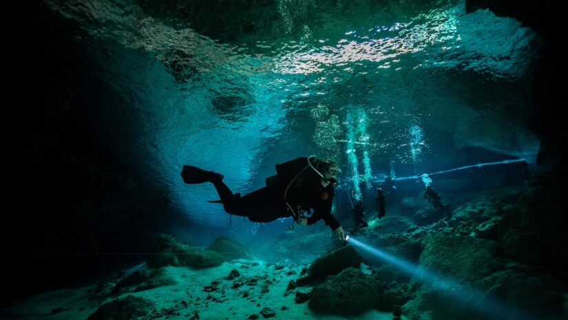 An underwater photograph of a scuba diver exploring a cave or cenote. The diver is swimming horizontally through clear turquoise water, shining a bright dive light onto the rocky floor. Several other divers, bubbles, and a straight diving guideline are visible in the background beneath the water's reflective surface.