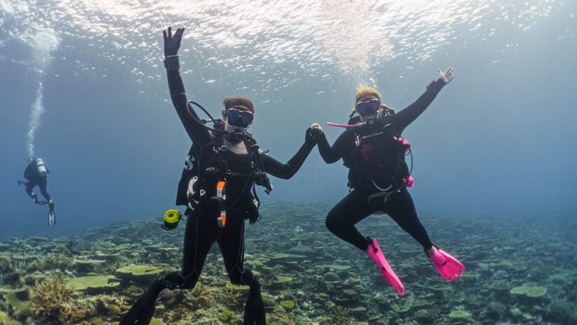 Two female scuba divers holding hands and cheering with their arms raised underwater over a vibrant coral reef in Fiji