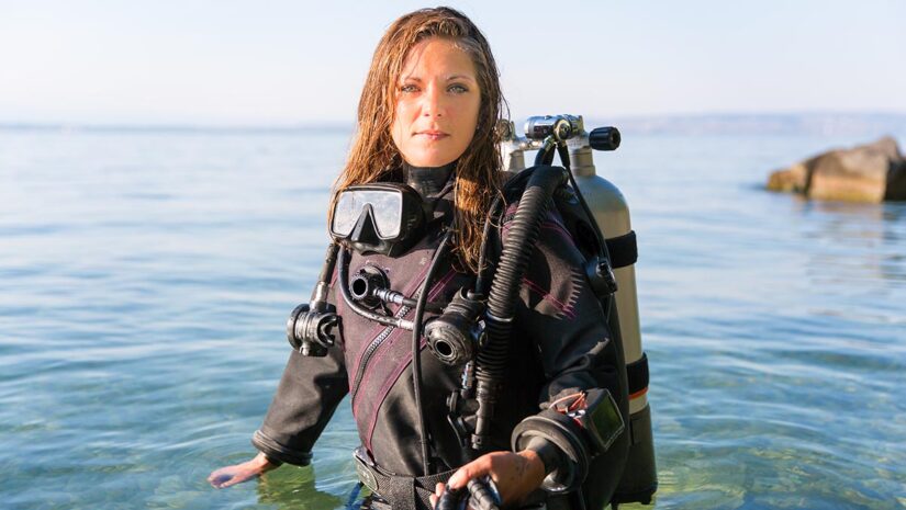 Female Scuba Diving Instructor Standing in Water Wearing a DrySuit, a Twin Tank and Holding Fins