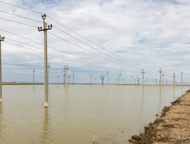 A rural area is flooded, with power lines rising above the water level under a cloudy sky, indicating recent heavy rainfall.