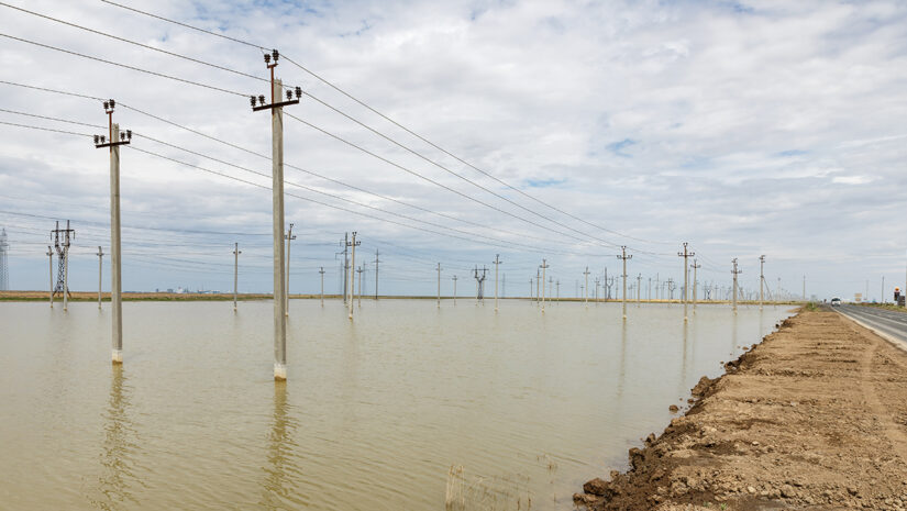 A rural area is flooded, with power lines rising above the water level under a cloudy sky, indicating recent heavy rainfall.