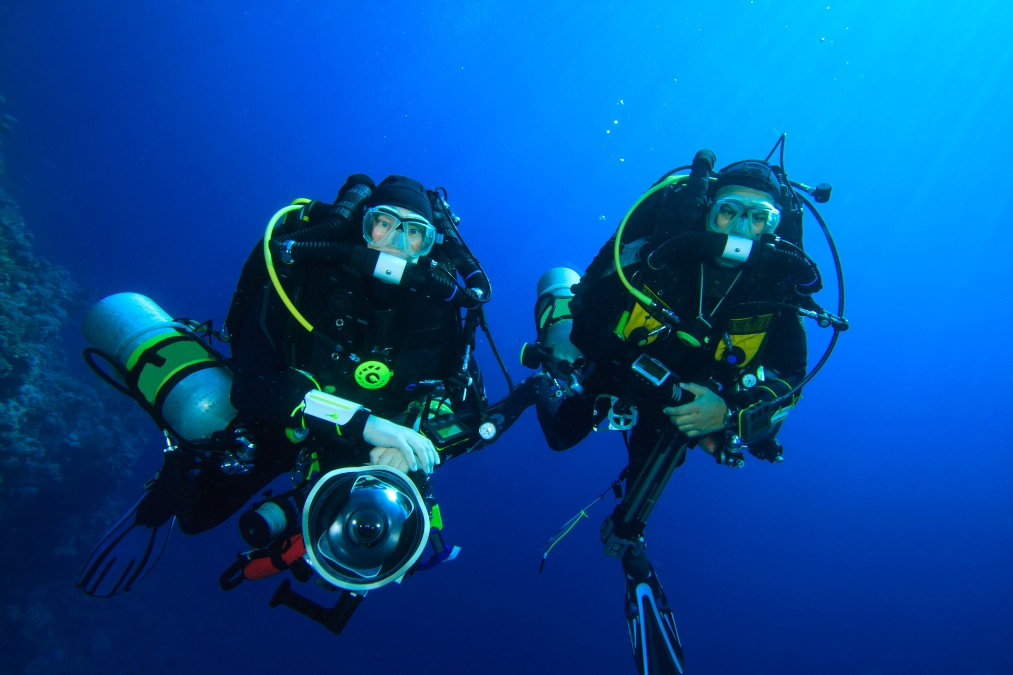 two fully equipped divers pose in front of camera with their arms out
