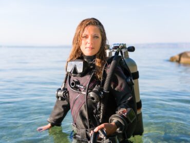 female scuba diving instructor standing in water wearing a dry suit a twin tank and holding fins