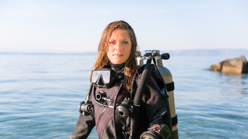 female scuba diving instructor standing in water wearing a dry suit a twin tank and holding fins