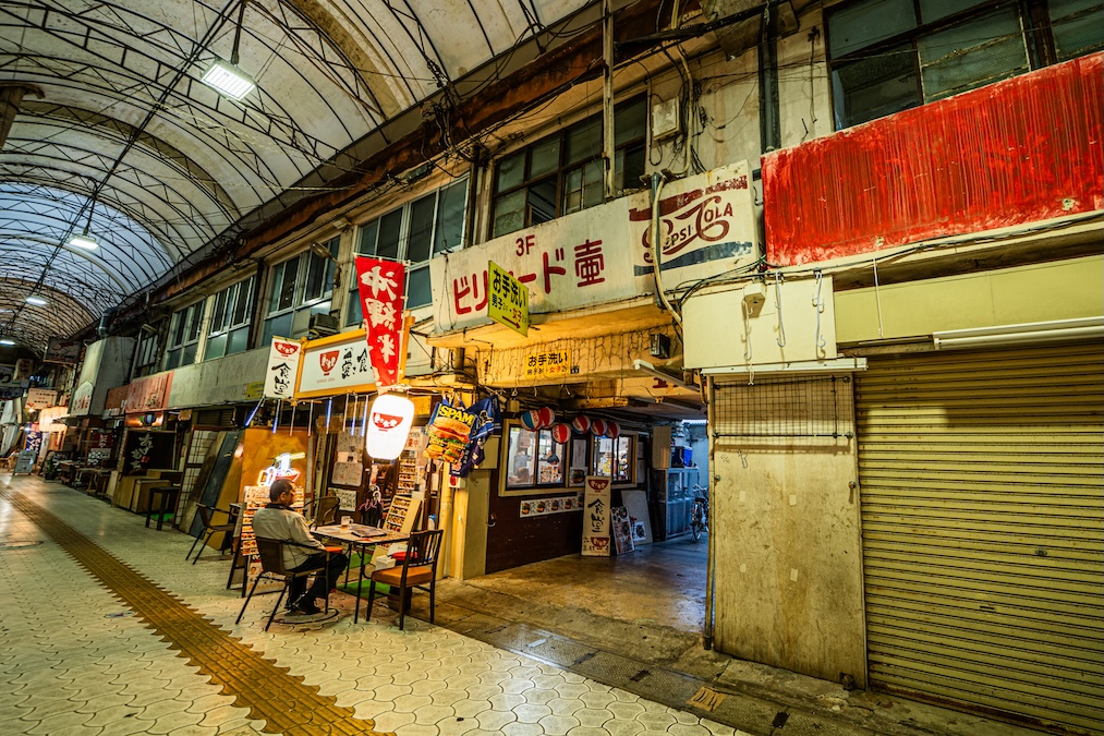 A quiet, covered shopping arcade in Okinawa featuring a small local eatery with a man sitting at an outdoor table, beneath a faded vintage Pepsi-Cola sign