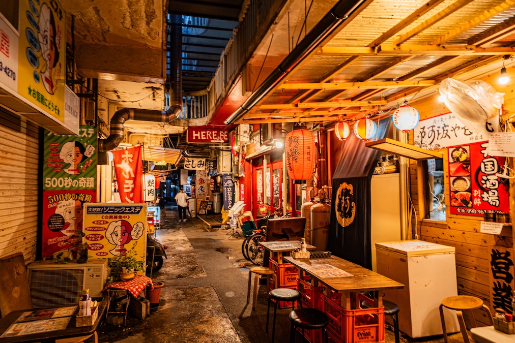 A vibrant, narrow alleyway at night in Okinawa, lined with small izakaya restaurants illuminated by warm paper lanterns, featuring outdoor seating made from stacked crates