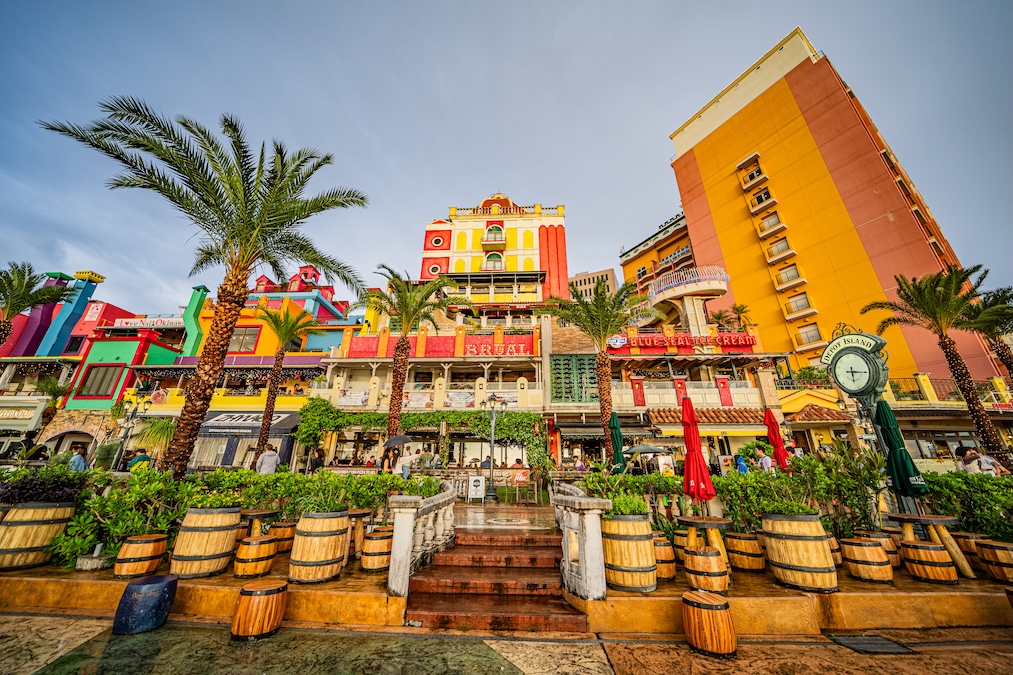 Vibrant, multi-story buildings painted in bright yellow, orange, red, and blue at Depot Island in Okinawa, featuring palm trees, a Blue Seal Ice Cream sign, and an outdoor seating area with tables and stools made from wooden barrels
