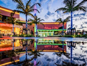 A colorful sunset view of the Mihama 7 Plex theater in Okinawa's American Village, featuring neon lights and palm trees perfectly reflected in a large water puddle on the ground