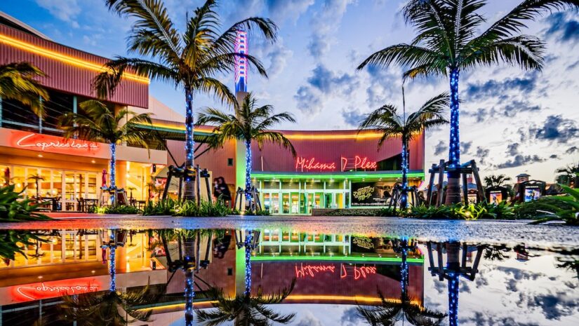 A colorful sunset view of the Mihama 7 Plex theater in Okinawa's American Village, featuring neon lights and palm trees perfectly reflected in a large water puddle on the ground
