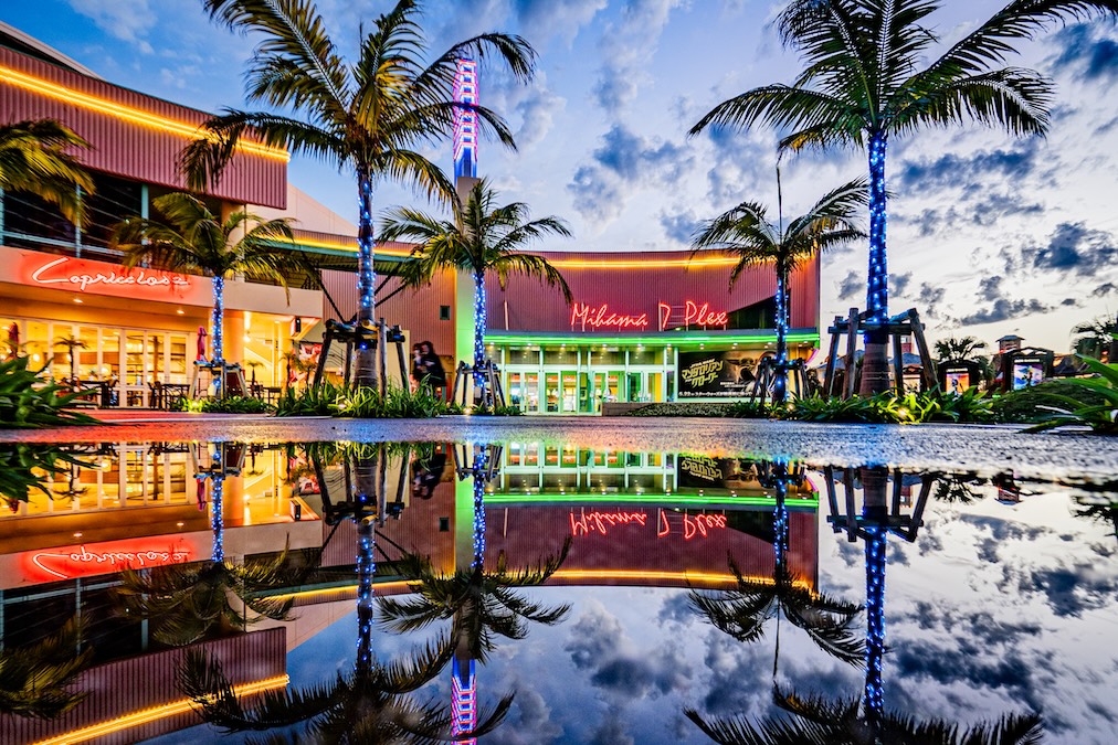 A colorful sunset view of the Mihama 7 Plex theater in Okinawa's American Village, featuring neon lights and palm trees perfectly reflected in a large water puddle on the ground