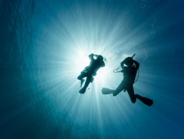 An underwater silhouette of two scuba divers, dramatically backlit by bright sunrays piercing down through the ocean surface