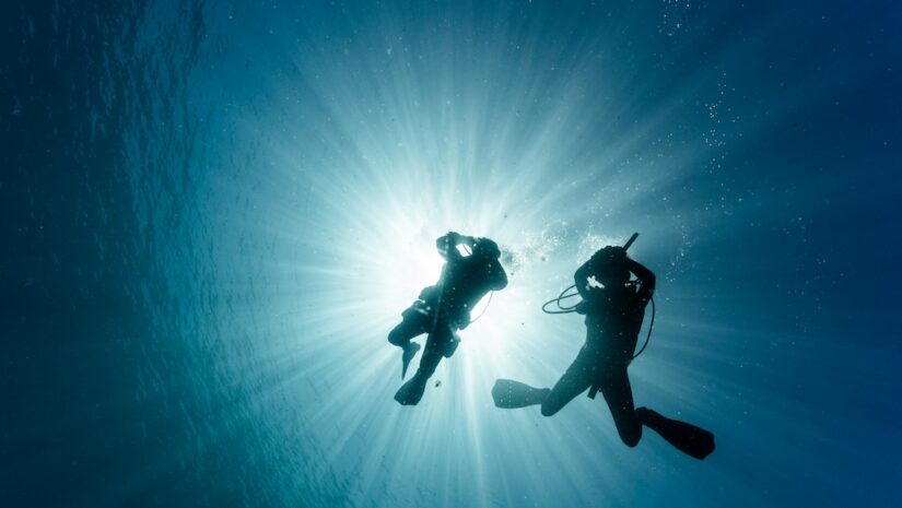 An underwater silhouette of two scuba divers, dramatically backlit by bright sunrays piercing down through the ocean surface