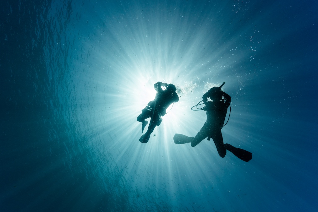 An underwater silhouette of two scuba divers, dramatically backlit by bright sunrays piercing down through the ocean surface