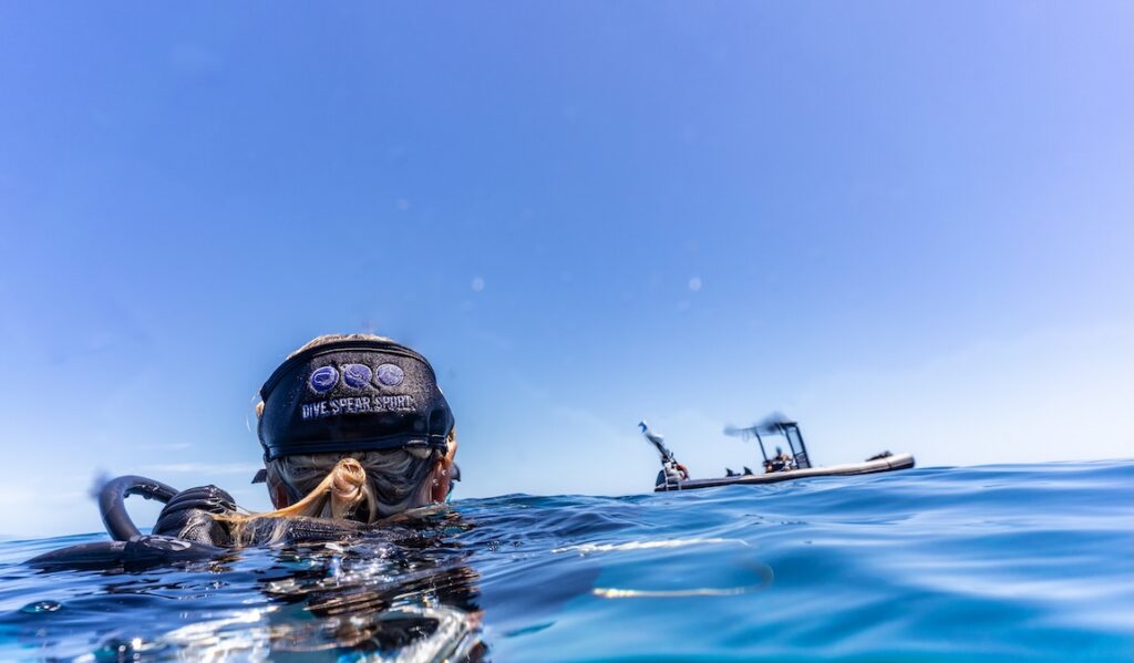A water-level view of the back of a diver's head at the surface, wearing a black "DIVE SPEAR SPORT" headband, with a small dive boat visible in the background under a clear blue sky