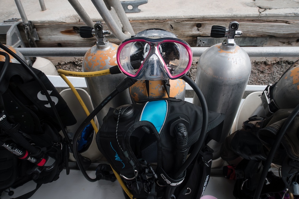 Close-up of assembled scuba diving equipment resting on a boat deck, featuring an aluminum tank, a black buoyancy compensator device (BCD) with blue accents, and a clear diving mask with a bright pink frame hanging over the regulator.