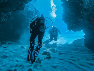 Two scuba divers swimming through a vibrant blue underwater cave or rock channel. The diver in the foreground is viewed from behind, wearing black gear and blue-accented fins, while the second diver follows closely behind them over a rippled sandy bottom