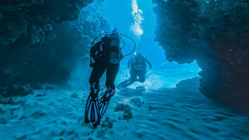 Two scuba divers swimming through a vibrant blue underwater cave or rock channel. The diver in the foreground is viewed from behind, wearing black gear and blue-accented fins, while the second diver follows closely behind them over a rippled sandy bottom