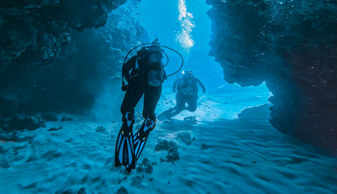 Two scuba divers swimming through a vibrant blue underwater cave or rock channel. The diver in the foreground is viewed from behind, wearing black gear and blue-accented fins, while the second diver follows closely behind them over a rippled sandy bottom
