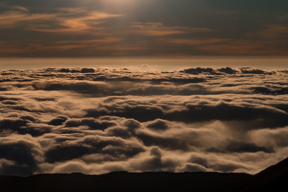 A high-altitude, dramatic view looking out over a thick, textured blanket of clouds illuminated by the warm, golden light of a sunrise or sunset. A dark, silhouetted landmass is visible at the very bottom edge of the frame