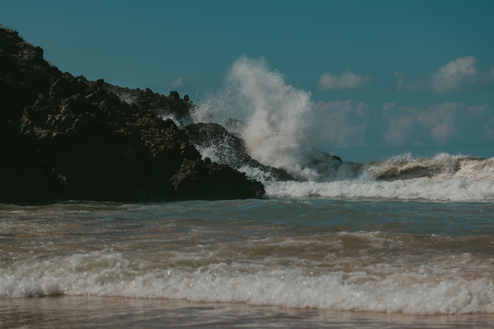 Powerful white ocean waves crashing violently against a rugged, dark rocky cliffside under a partially cloudy, deep blue sky