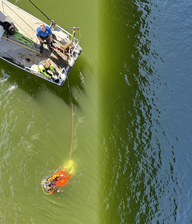 Austin PD diver fully equipped in a high-visibility orange drysuit holding swim fins near a body of water