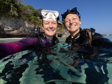 Two smiling women in wetsuits and scuba gear taking a selfie while partially submerged in clear, shallow ocean water. Both have diving masks resting on their foreheads, with a rocky coastline and trees in the background