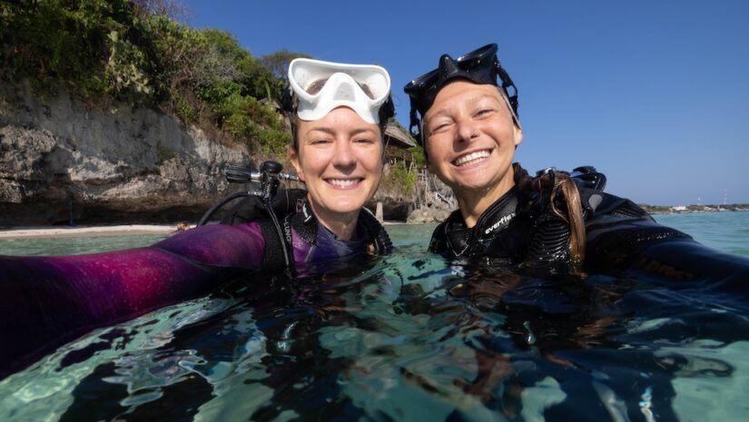 Two smiling women in wetsuits and scuba gear taking a selfie while partially submerged in clear, shallow ocean water. Both have diving masks resting on their foreheads, with a rocky coastline and trees in the background