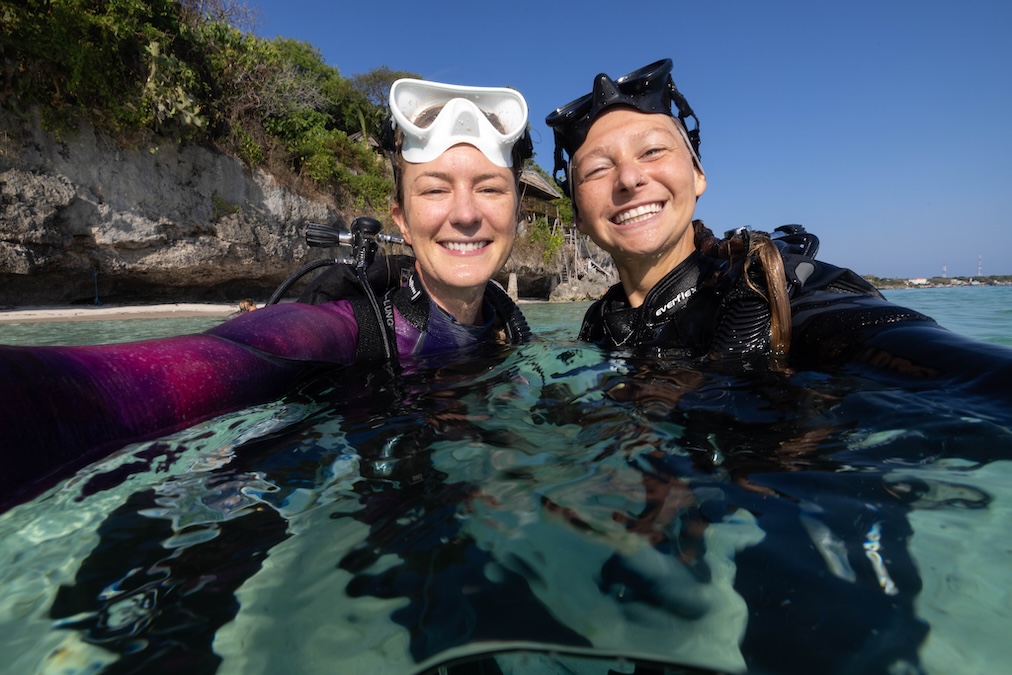 Two smiling women in wetsuits and scuba gear taking a selfie while partially submerged in clear, shallow ocean water. Both have diving masks resting on their foreheads, with a rocky coastline and trees in the background