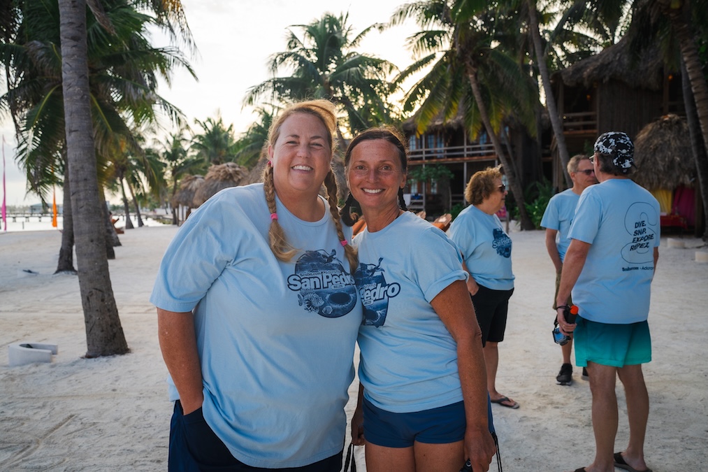 Two women posing together and smiling on a tropical, sandy beach lined with palm trees. Both are wearing matching light blue "San Pedro Belize" t-shirts, with thatched-roof structures and other people in the background