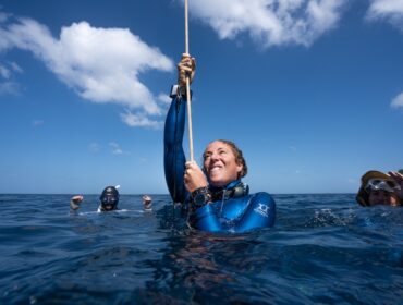 A smiling woman in a blue wetsuit surfaces from deep blue water, gripping a rope with both hands. She is looking upward. In the background at water level, two other divers are visible. The sky above is bright blue with scattered white clouds
